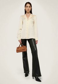 Kremowa koszula z długim rękawem, kołnierzykiem, guziczkami z przodu i subtelnymi złotymi akcentami, zestawiona z czarnymi, rozkloszowanymi skórzanymi spodniami i brązową torebką.