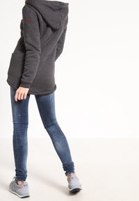 Mörkgrå fleecetröja med huva, långa ärmar och sidofickor. Buren över blåa skinny jeans och gråa sneakers.