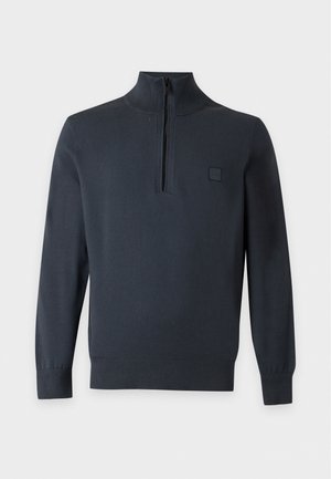 Jeune homme portant un pull noir zippé par-dessus une chemise à carreaux, un short bleu marine, des chaussettes blanches et des mocassins noirs, debout devant un fond uni.