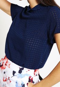 Mörkblå topp med kort ärm i halvgenomskinligt tyg med rutmönster, som har en subtil krage och draperad halsringning.