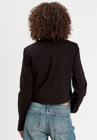 Black cropped jacket with long sleeves and a structured design. Worn over light blue high-waisted jeans, showcasing a casual look.