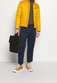 Gul puffjacka med dragkedja och ståkrage, olivgrön tröja, blå byxor, bruna sneakers och en svart ryggsäck med klafföppning.