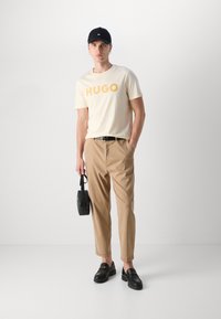T-shirt beige clair avec "HUGO" en jaune, chinos beige, mocassins noirs avec des accents dorés, et une casquette de baseball noire, tenant un petit sac noir.