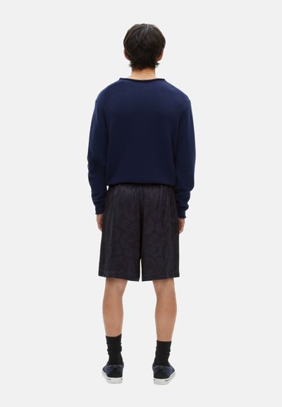 Marineblaues Strickoberteil, schwarze gemusterte Shorts, schwarze Socken und dunkle Schuhe. Sichtbare Nähte und lockere Passform. Minimalistische Ästhetik.