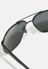 Ochelari de soare aviator negri cu lentile închise, cadru metalic, brațe subțiri și suporturi de nas transparente. Prezentare cu un design minimalist.