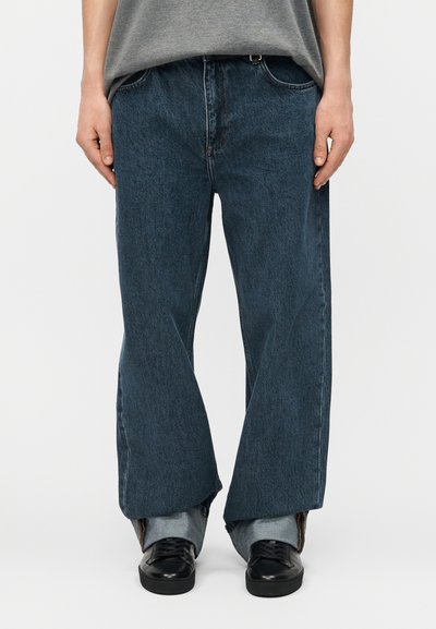 Persona vistiendo vaqueros sueltos de mezclilla azul oscuro con dobladillo en el extremo, camisa gris y zapatos de cuero negros sobre un fondo liso.