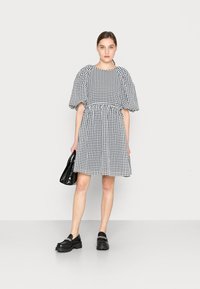 Vestido preto e branco em padrão xadrez, com mangas bufantes, cintura ajustada, saia até ao joelho, combinado com sapatos pretos e uma pequena mala.