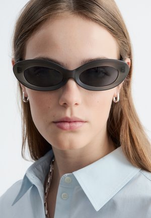 Joven mujer con largo cabello castaño claro lleva gafas de sol ovaladas negras, pendientes de aro plateados, una camisa azul claro abotonada y un collar.
