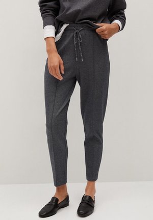Person iført mørkegrå, tilspidsede bukser med snøre i taljen, kombineret med sorte slip-on sko og en mørk langærmet top.