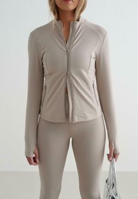 Beige Sportjacke mit Reißverschluss, langen Ärmeln und Seitentaschen, aus dehnbarem Material. Kombiniert mit passenden Leggings. Silberne Tasche in der Hand.