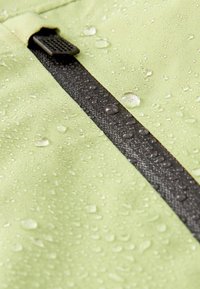 Hellgrüner wasserdichter Stoff mit Wassertropfen und einem geschlossenen schwarzen Reißverschluss mit strukturiertem Ziehgriff.
