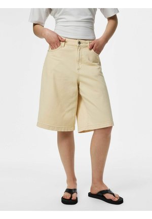 Persoană care poartă pantaloni scurți bej până la genunchi, top alb și sandale negre, stând cu mâinile în buzunare pe un fundal alb.