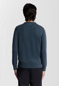 Mörkblå tröja för män med rundad hals, texturerad stickad tyg, ribbade manschetter och fåll, etikett vid nacken, figurnära design. Visar baksidan.