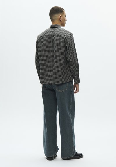 Kariirana košulja tamnih tonova, opuštenog kroja, uparena s širokim plavim trapericama. Sadrži ovratnik i duge rukave. Jednostavan dizajn.