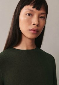 Dunkelgrüner gerippter Pullover mit rundem Halsausschnitt, getragen von einem Modell mit glattem schwarzen Haar. Glatte Textur, minimalistische Details.