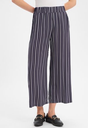 Vrouw die marineblauwe wijde broek met witte verticale strepen en zwarte loafers draagt, staand tegen een effen witte achtergrond.