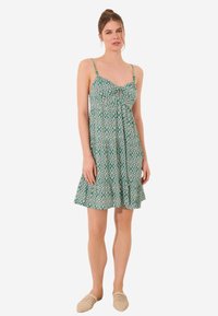 Robe verte à motifs avec fines bretelles, présentant un buste froncé et une jupe évasée avec un léger volant à l'ourlet. Tissu léger.