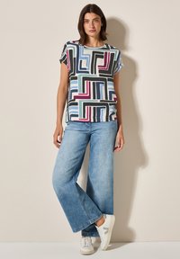 Un haut à manches courtes à motif géométrique multicolore associé à un jean bleu délavé à jambes larges et des baskets blanches avec des accents gris.