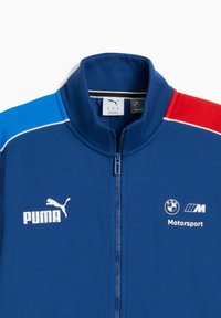 Blå zip-up jakke med Puma- og BMW Motorsport-logoer, og røde og lyseblå accenter på skuldrene.