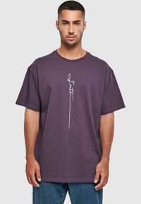 Kortærmet t-shirt med rund hals i mørk lilla med tekstureret stof og hvidt skriftdesign på forsiden, sammen med blå denim jeans.