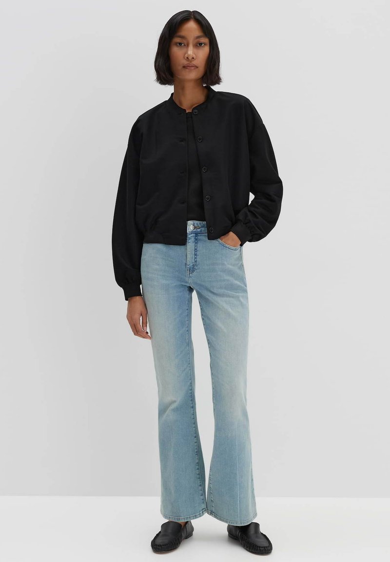 Schwarze Bomberjacke mit rundem Ausschnitt und Knopfverschluss, kombiniert mit hellblauen Schlagjeans und schwarzen Loafers. Einfaches Design, lässige Passform.