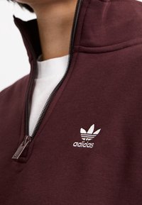 Mörkröd zip-up sweatshirt tillverkad av texturerad tyg, med en vit broderad Adidas-logotyp på bröstet och en silverdragkedja.