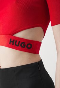Top corto rojo acanalado con una banda negra ancha que presenta el texto "HUGO", mostrando piel a través de un diseño con cortes. Combinado con pantalones negros de tiro alto.