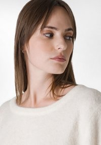 Weißer Strickpullover mit rundem Ausschnitt. Weiche Textur, lässige Passform. Die Haare sind lang und glatt, leicht zur Seite gescheitelt. Neutraler Hintergrund.