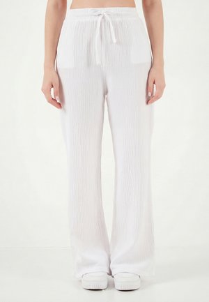 Kvinde iført løstsiddende, hvide teksturerede bukser med snørre og hvide sneakers, stående mod en ensartet lys baggrund.