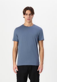 Jauns vīrietis stāv, tērpies vienkrāsainā zilā īsām piedurknēm T-kreklā un melnās biksēs, uz vienkārša baltā fona.