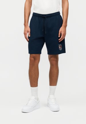 Man draagt marineblauwe shorts met trekkoord en Tommy Hilfiger-logo, witte sokken en witte sneakers tegen een effen lichte achtergrond.