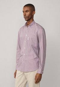 Camicia a manica lunga a quadri rossi e bianchi con colletto button-down, caratterizzata da una tasca sul petto e bottoni bianchi, abbinata a pantaloni di colore chiaro.