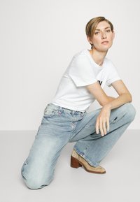 Light blue wide-leg jeans with a faded finish, paired with a fitted white t-shirt. The model wears tan heeled ankle boots. Simple, clean design.