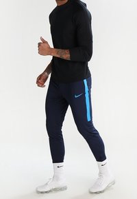 Czarna, długorękawowa koszulka sportowa w połączeniu z granatowymi dresami z niebieskim paskiem i logo. Uzupełniona białymi skarpetkami i butami na przezroczystej podeszwie.