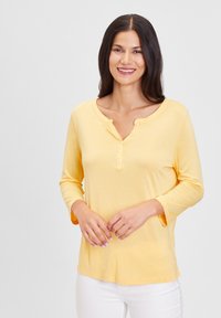 Gelber Langarmoberteil mit geknöpftem Ausschnitt, weichem Material und lässiger Passform, kombiniert mit weißen Hosen.