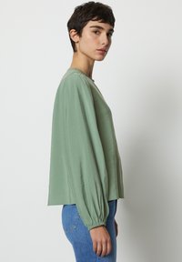 Blusa verde con maniche lunghe arricciate, scollo rotondo e vestibilità rilassata. Tessuto liscio con una leggera lucentezza, abbinata a jeans blu.