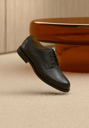 Chaussure formelle en cuir noir avec lacets reposant sur un tapis beige devant une table ronde marron brillante dans une pièce neutre.