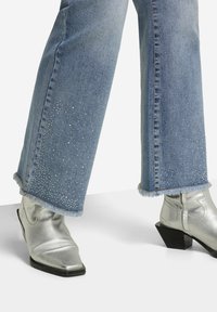 Ljust blå denim flare-jeans med fransade nederkanter, dekorerade med spridda rhinestones. Kombinerad med silverfärgade metallicankelstövlar med fyrkantig tådesign.