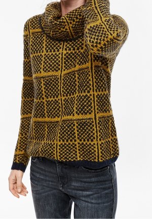 Femme portant un pull en tricot à carreaux jaune moutarde et noir avec un col bénitier et un jean foncé, main touchant le cou, cadré sous le nez.