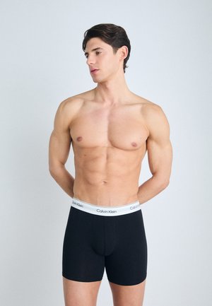 Fekete pamut boxer alsónadrág fehér elasztikus derékpánttal, amelyen a "Calvin Klein" logo látható; a modellen a kezében a háta mögött áll, minimalista háttérrel.