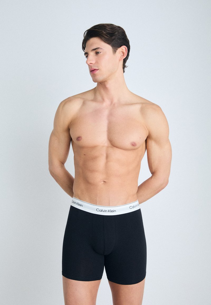 Crne pamučne bokserice s bijelim elastičnim pojasom na kojem se nalazi logo "Calvin Klein", model stoji s rukama iza leđa, minimalna pozadina.