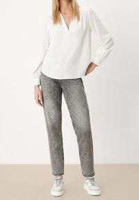 Donna che indossa una blusa bianca larga, jeans slim grigi sbiaditi e sneakers bianche con piattaforma, in piedi con una mano sulla vita.