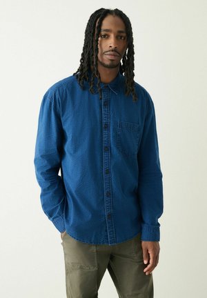 Mann mit langen Dreadlocks, der ein blaues Hemd mit Knöpfen und olivgrüne Hosen trägt, vor einem schlichten hellen Hintergrund stehend.