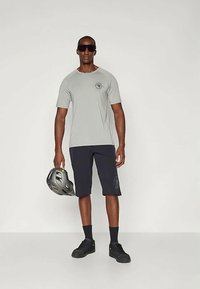 Muž v šedej tričku s krátkym rukávom, čiernych šortkách, čiernych ponožkách a topánkach, v ľavej ruke drží šedú cyklistickú prilbu a má na sebe veľké slnečné okuliare.