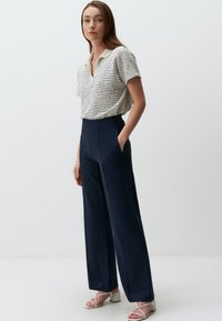 Biały teksturowany top z krótkim rękawem, w połączeniu z granatowymi spodniami z wysokim stanem i białymi sandałkami na obcasie. Strój ma dekolt w kształcie litery V oraz boczne kieszenie.