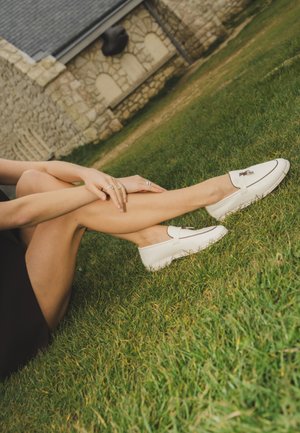 Kvinde iført hvide loafers siddende på græs, krydser benene, med stenhuset i baggrunden.