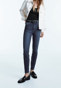 Tummansiniset korkeavyötäröiset farkut, joissa on slim fit -leikkaus, yhdistettynä mustaan toppiin ja vaaleansävyiseen takkiin, asusteena musta vyö ja loafertossut.