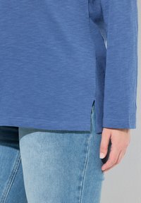 Modré tričko s dlouhým rukávem z měkké látky, s bočními rozparky na lemu a volným střihem, v kombinaci se světle modrými džínami.