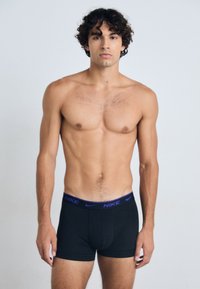 Schwarze Boxer-Brief mit einem lila Logo-Bund. Der Stoff wirkt glatt und elastisch, entworfen für Komfort und Passform. Das Modell steht aufrecht.