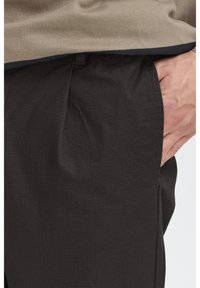 Pantalones negros con textura, pliegue frontal y bolsillos laterales, combinados con una camiseta beige que tiene un borde oscuro. Vista en primer plano del área del bolsillo.
